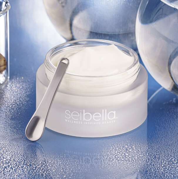 Sei Bella Advanced Epidermal Perfector Cream in an open white jar with a silver spatula, displayed on a reflective blue surface.